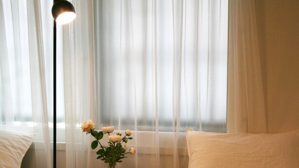 Une chambre à coucher avec un lit, une table de chevet ornée d’un vase de fleurs et des rideaux légers.