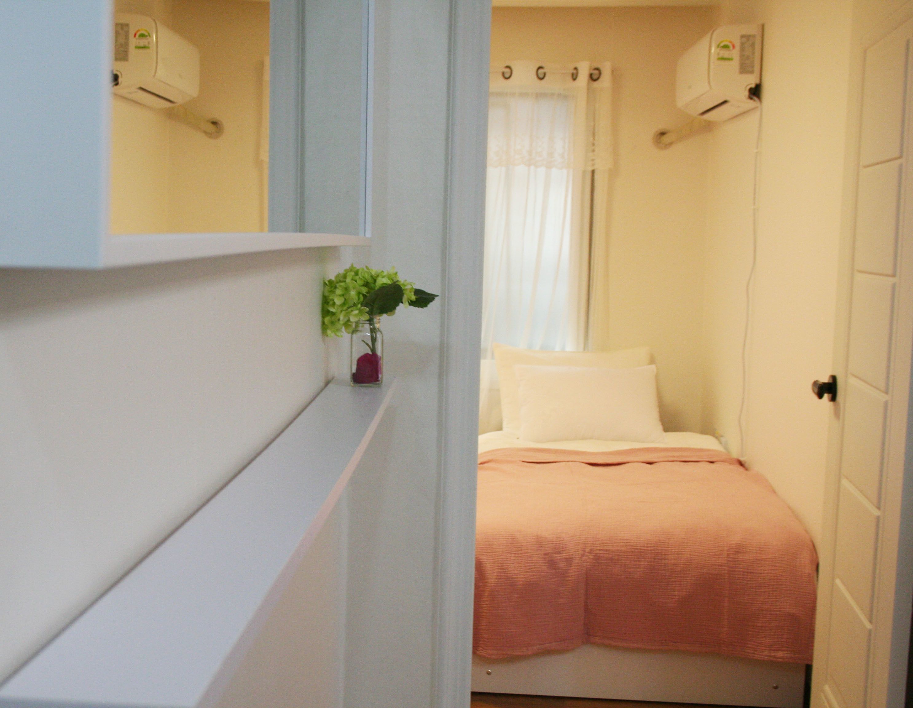 A small, narrow room with a bed, a shelf with a vase of flowers, and a door.