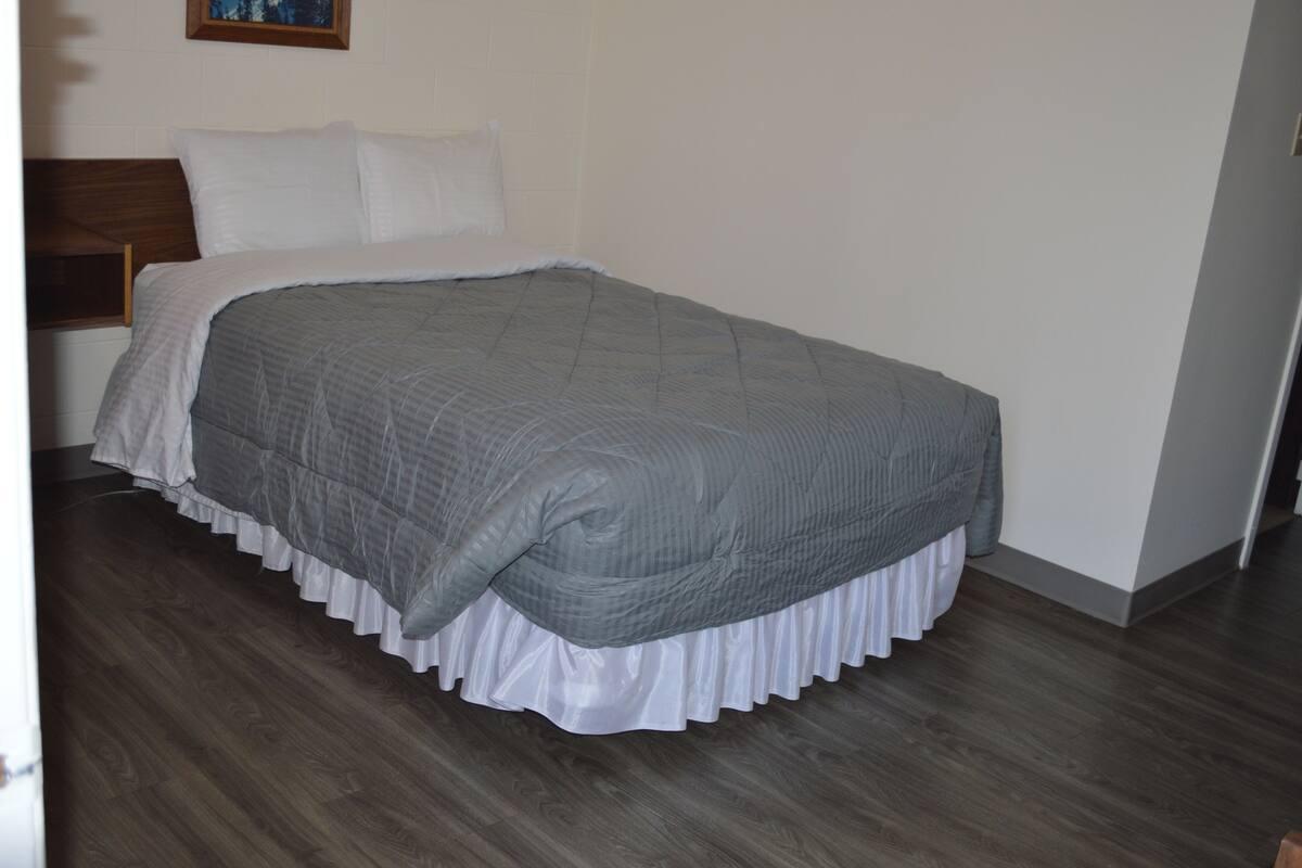 Una cama bien hecha con colcha gris y falda blanca, cabecera de madera y pared blanca con un cuadro enmarcado arriba.
