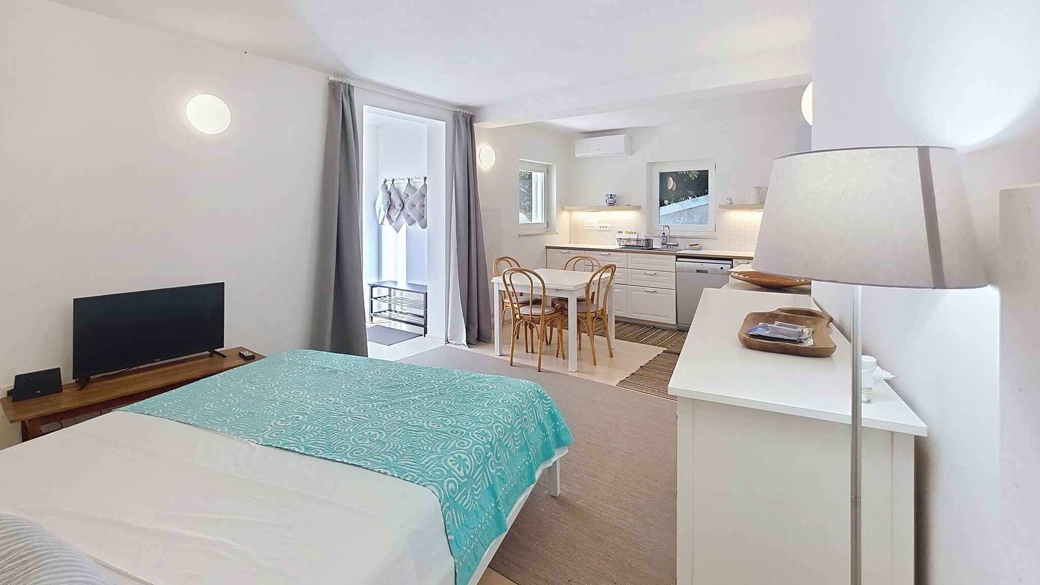 A hotel room with a bed, a desk with a lamp, a TV, a balcony with a view, and a kitchenette.
