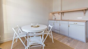 A white dining table with four chairs, a small tray with glasses, and a light-colored kitchen with white cabinets and a wooden countertop. - 2-room apt. 32sqm - lodging house Rosa-Lena (Norderney)
