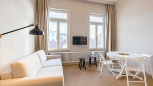 A modern living room with a white sofa, a round dining table with chairs, a small black table, and a TV mounted on the wall. - 2-room apt. 32sqm - lodging house Rosa-Lena (Norderney)