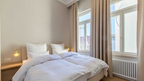 A hotel room with a large bed, a nightstand with a lamp, a window with curtains, and a radiator. - 2-room apt. 32sqm - lodging house Rosa-Lena (Norderney)