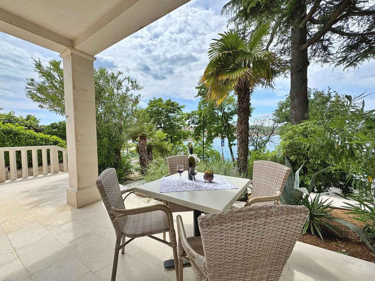 A covered outdoor seating area with wicker chairs and a table, surrounded by lush greenery and a view of the sea.