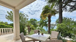 A covered outdoor seating area with wicker chairs and a table, surrounded by lush greenery and a view of the sea.