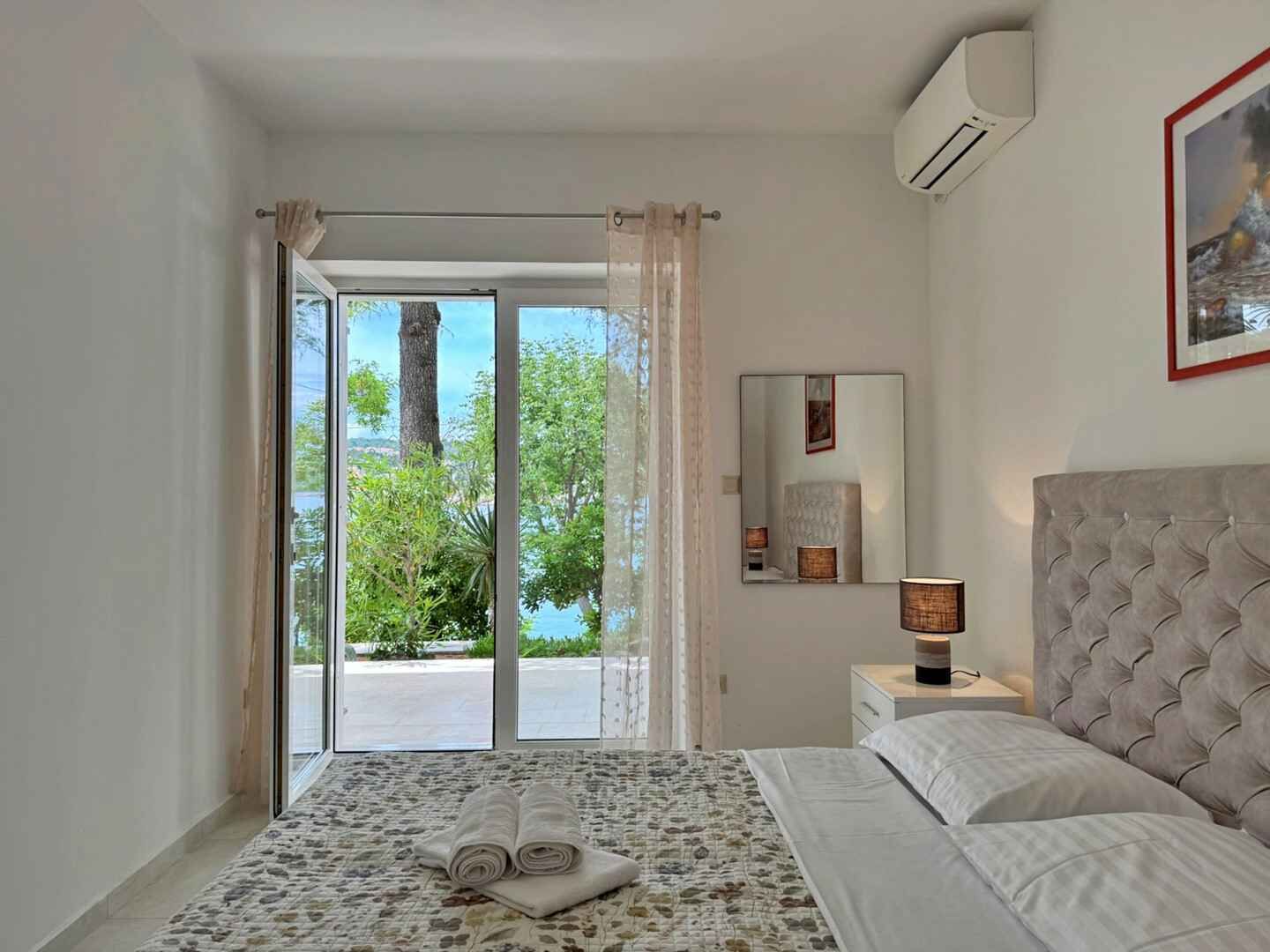 A bedroom with a bed, a nightstand, a lamp, a mirror, and a view of the outdoors.