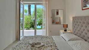 Quarto com cama, mesa de cabeceira, abajur, espelho e vista para o exterior.