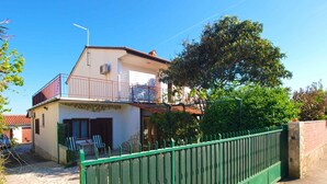 Ein zweigeschossiges Haus mit Balkon, Steinfassade und grünem Metallzaun.