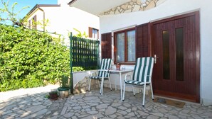 Eine Terrasse im Freien mit Steinboden, zwei gestreifte Stühle, einem Tisch und Blick auf ein weißes Gebäude mit Steinvorsatzfassade und Holzläden.