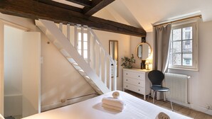 Une chambre à coucher avec un lit, une chaise, une commode, un miroir et une fenêtre avec des rideaux.