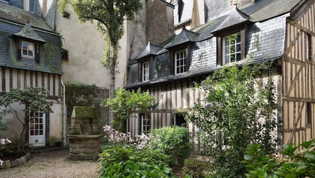 Une cour bordée de maisons traditionnelles à colombages, avec une fontaine en pierre et des plantes en fleurs.