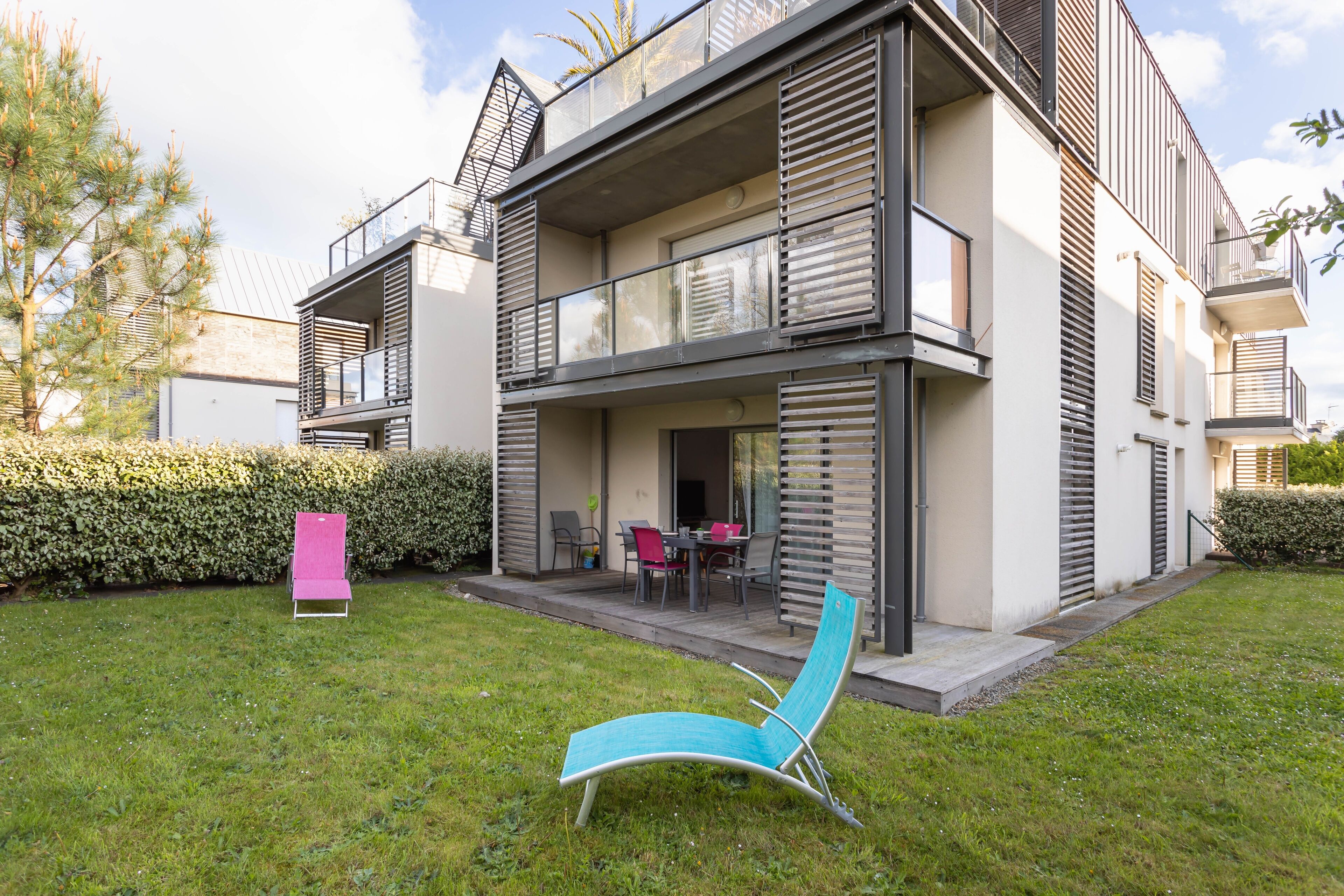 En modern bostadsbyggnad i två våningar med en balkong, en uteplats med utemöbler och en välskött gräsmatta.