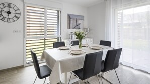 A dining area with a table set for four, a clock on the wall, and a window with blinds. - LE GARDEN - Vaste appartement avec Jardin et terrasse (Dinard)