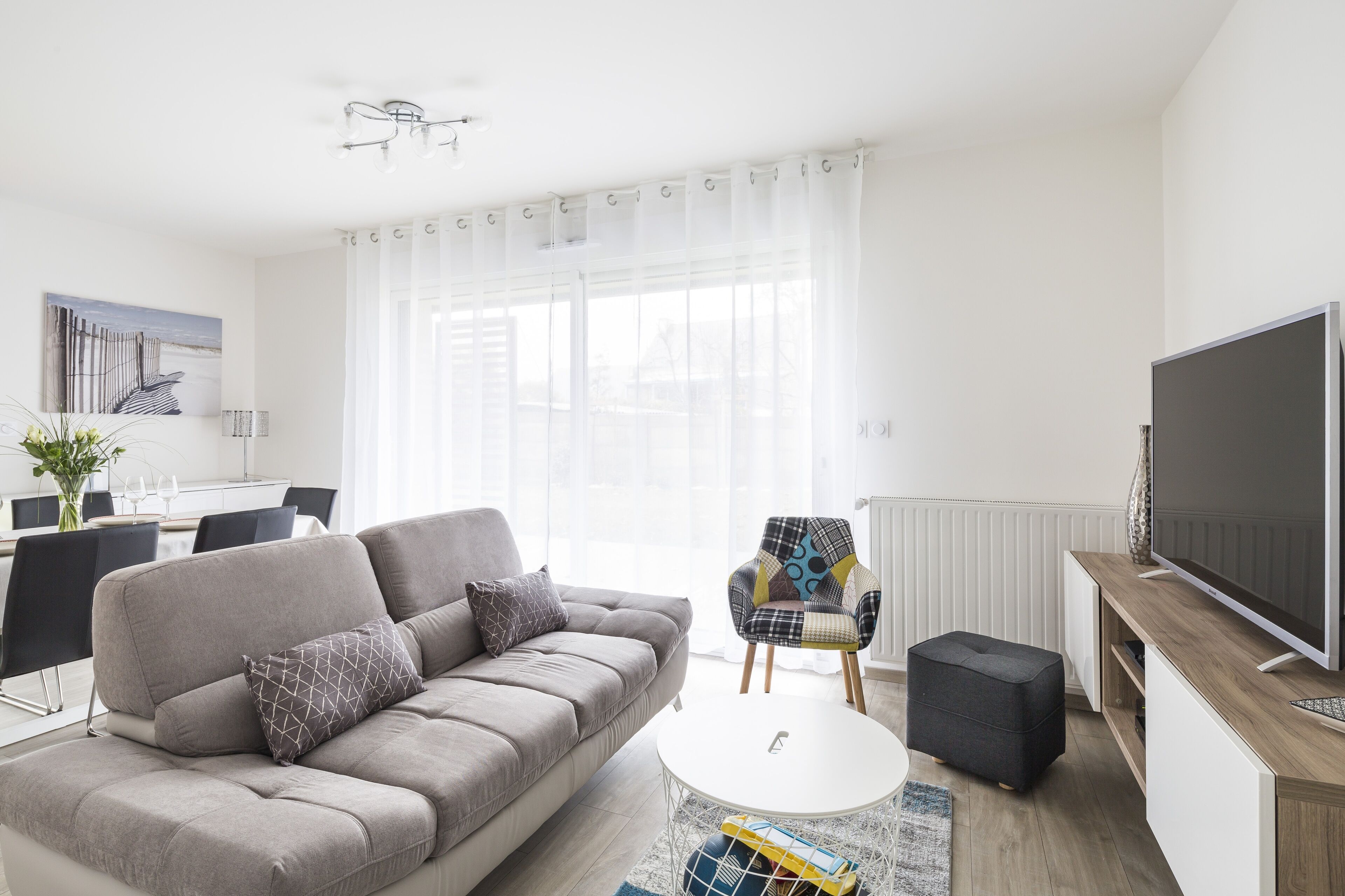 Un salon moderne avec un canapé gris, un fauteuil à motifs, un téléviseur à écran plat et un coin repas à l’arrière-plan.