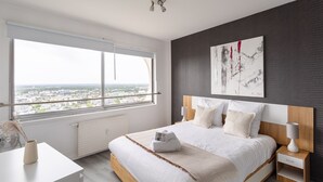 Un dormitorio moderno con un ventanal grande, una cama con ropa de cama blanca, una mesita de noche y una obra de arte colgada en la pared.