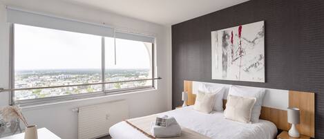 A modern bedroom with a large window, a bed with white linens, a bedside table, and a wall-mounted artwork.