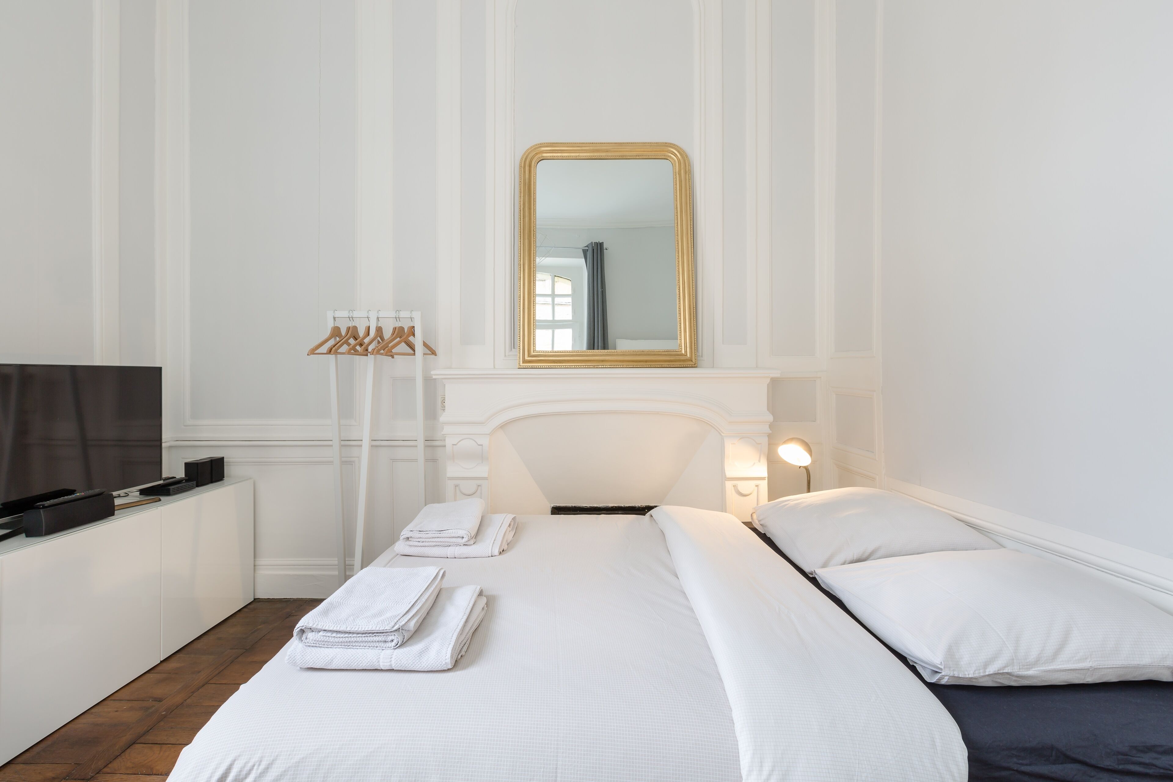 Une chambre d’hôtel avec un lit bien fait, un téléviseur posé sur un meuble, un miroir au-dessus du lit et un porte-manteau avec des cintres.