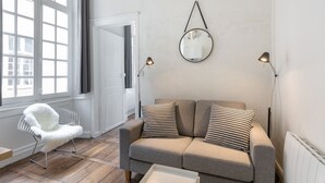 A modern living room with a grey sofa, two patterned cushions, a white chair, and a round mirror on the wall. - L'HORLOGE - Cocon de Charme au coeur de Rennes (Rennes)