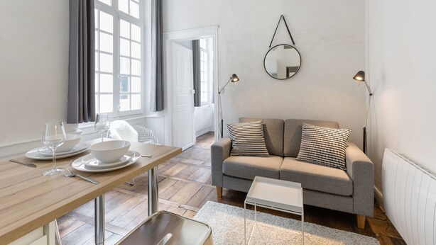 A modern living room with a sofa, a dining table set with plates and glasses, a mirror on the wall, and a window with curtains. - L'HORLOGE - Cocon de Charme au coeur de Rennes (Rennes)