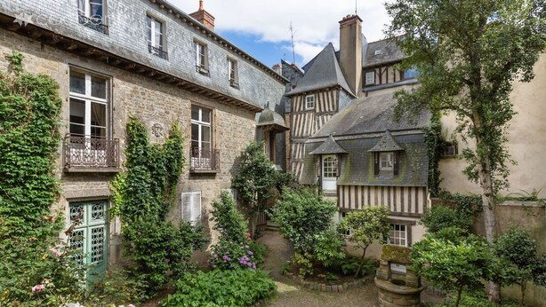 Une cour bordée de bâtiments en pierre, avec un puits en pierre et une végétation luxuriante.
