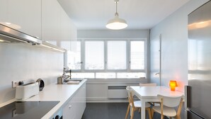 A modern kitchen with white cabinetry, a built-in oven, a dining table with chairs, and a window with blinds. - TREGOR - 2 ch - Parking + Vue sublime (Rennes)