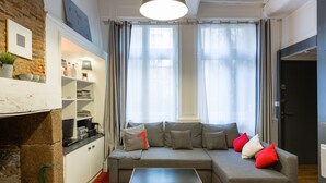 A modern living room with a grey sofa, red and white cushions, a small coffee table, and a red carpet. There is a fireplace on the left, a bookshelf, and a door on the right. - COCON URBAIN Centre Historique (Rennes)
