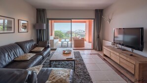 A modern living room with a leather sofa, a coffee table, a TV on a wooden stand, and a balcony with a view.