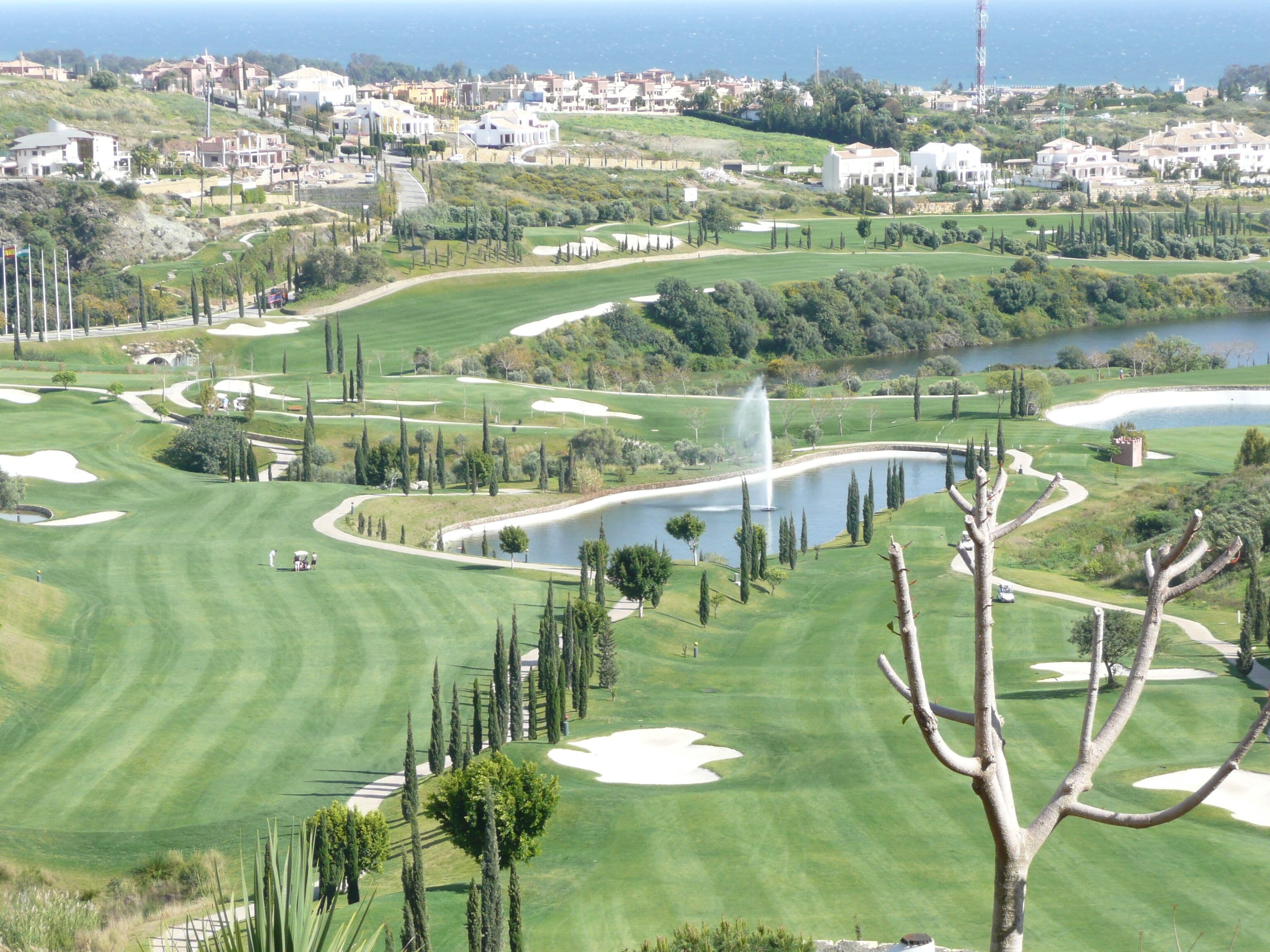 A golf course with bunkers, a water feature, and a view of a coastal town.