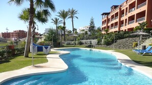 A swimming pool with a curved edge, surrounded by a grassy area with palm trees. There are lounge chairs and a building with balconies in the background.