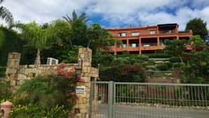 A multi-story building with balconies, surrounded by lush greenery and a stone wall.