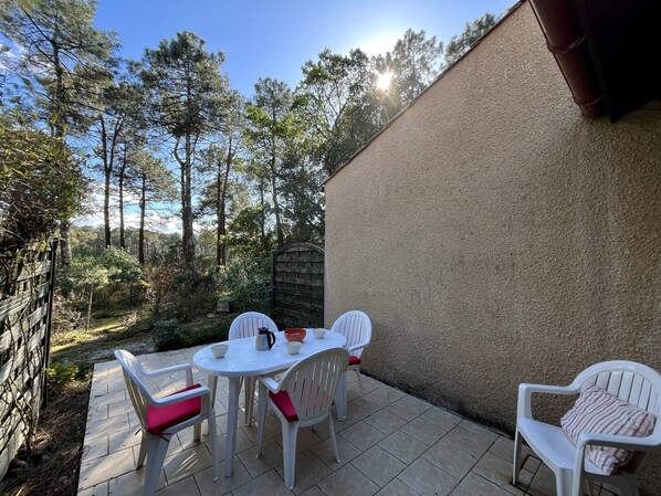 Une terrasse extérieure avec une table ronde et des chaises en plastique blanc, entourée d’une clôture en bois et d’arbres.