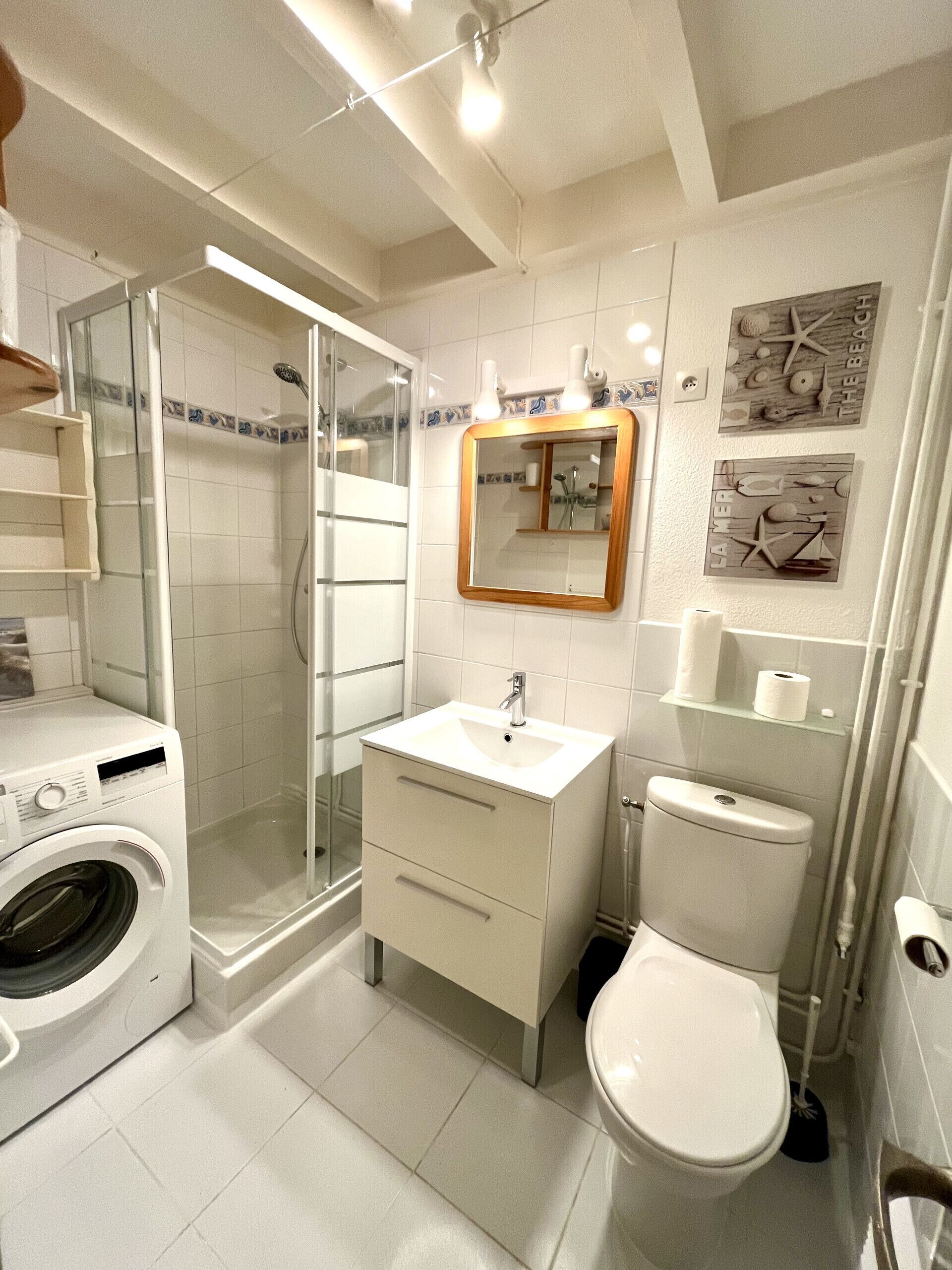 A compact bathroom with a glass shower enclosure, a built-in washing machine, a sink, and a toilet.