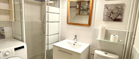 A compact bathroom with a glass shower enclosure, a built-in washing machine, a sink, and a toilet.