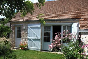 A rustic stone house with a tiled roof, a wooden door, and a small porch area with pink flowers.