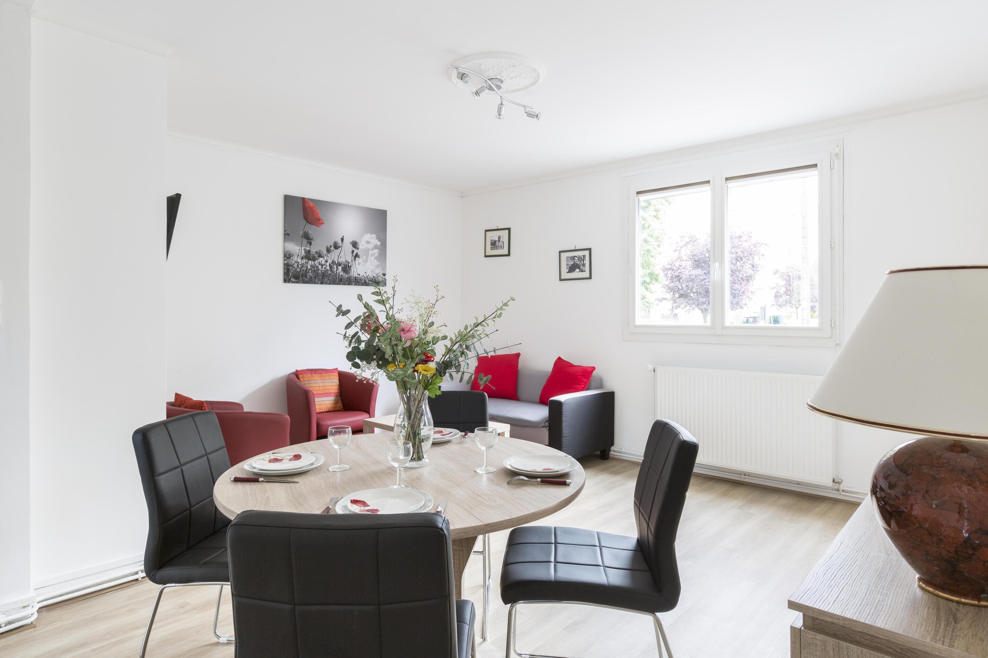 Un coin repas avec une table ronde, des chaises noires, un vase de fleurs et une lampe posée sur une petite table d’appoint.