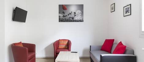 A living room with a red armchair, a grey sofa with red cushions, a small wooden table, and a red rug.