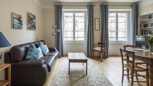 En stue med sofa, sofabord, spisebord med stole, bogreol og store vinduer med gardiner.