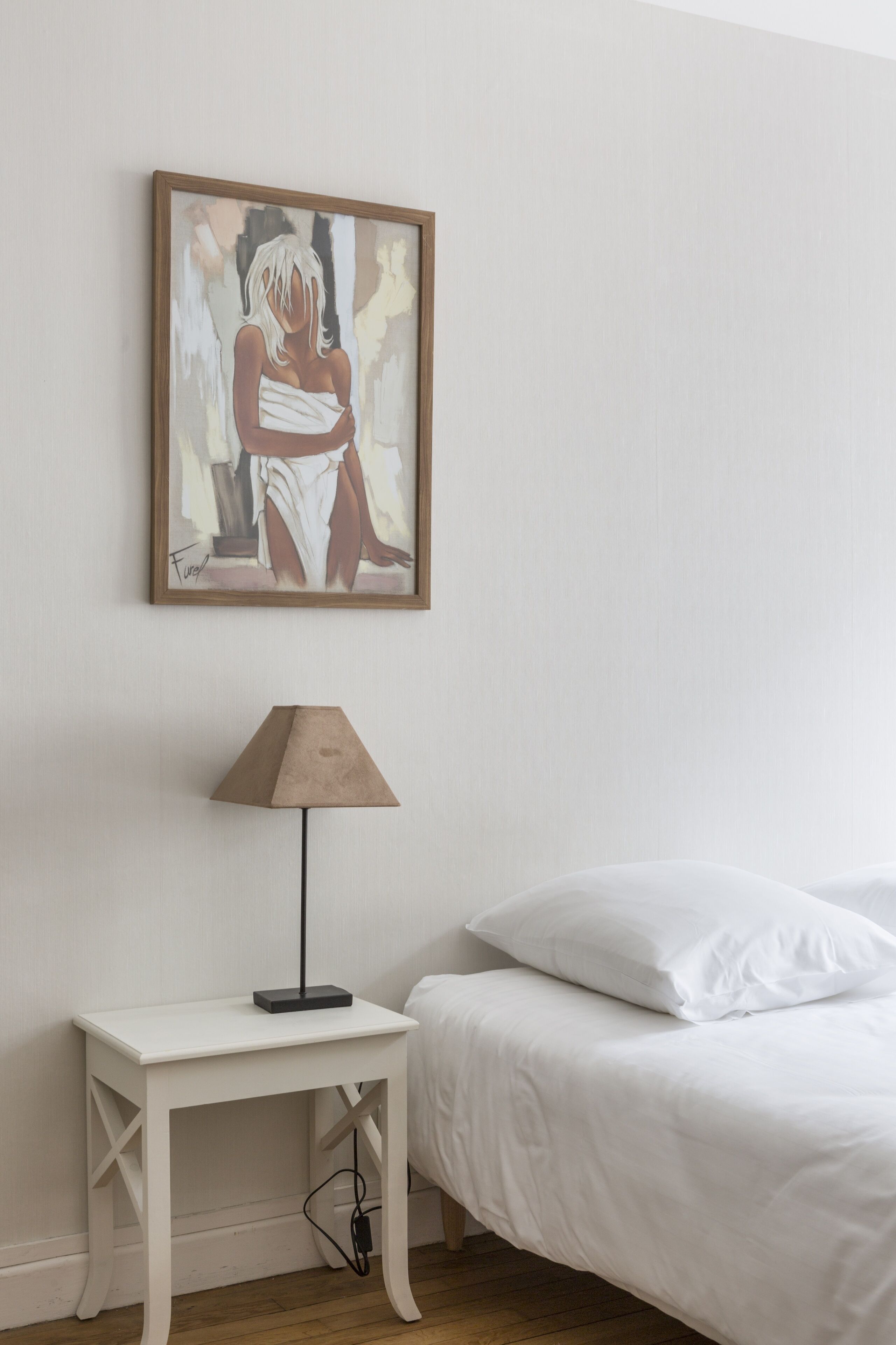 A bedroom with a white bed, a bedside table with a lamp, and a framed painting on the wall.