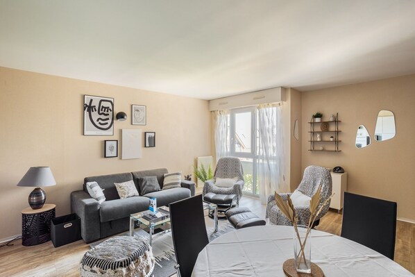Un salon moderne avec un canapé, une table basse et des fauteuils. On y trouve une table à manger ornée d’un vase contenant des plantes séchées. La pièce est dotée d’une grande fenêtre avec des rideaux et offre une vue sur l’extérieur.