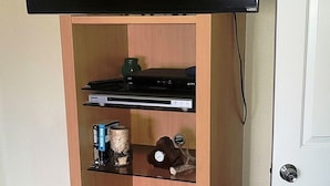 Une pièce avec un téléviseur fixé au mur, un meuble en bois avec des étagères, une table et une chaise.