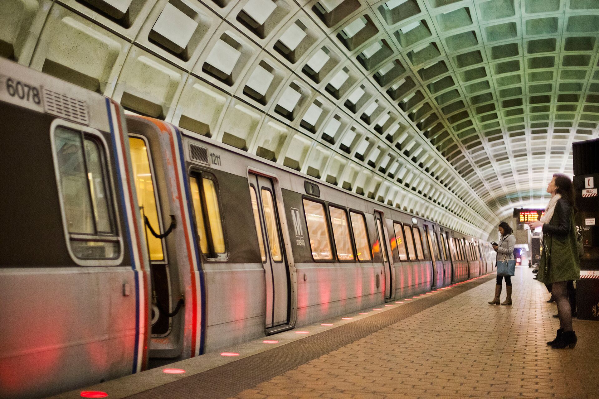 A subway station with a train at the platform, overhead lighting, and a tiled floor.