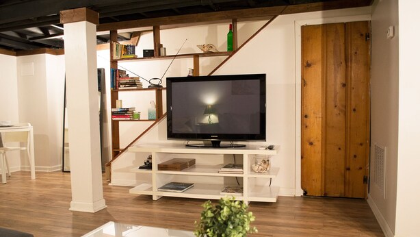 Ein modernes Wohnzimmer mit einem Flachbildfernseher auf einem weißen Schrank, Holzregalen mit Büchern und Dekorationsgegenständen, einem Glastisch mit einer Pflanze und einer Holztür im Hintergrund.
