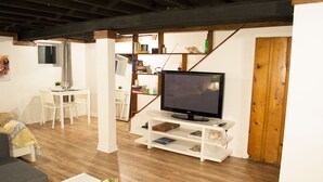 Ein Wohnzimmer mit Fernseher auf einem weißen Schrank, Holzboden, einem Essbereich mit weißen Stühlen und einer Treppe, die nach oben führt.