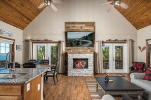 A spacious living area with a stone fireplace, wooden ceiling, and a large TV mounted above it.