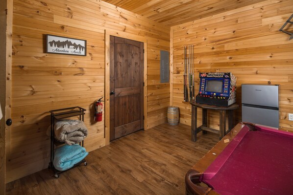 Interior de una cabaña de madera con mesa de billar, una máquina de pinball Midway, refrigerador y extintor.