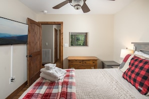 A bedroom with a bed, a wooden dresser, a nightstand, a ceiling fan, and a window with a view of the outdoors.