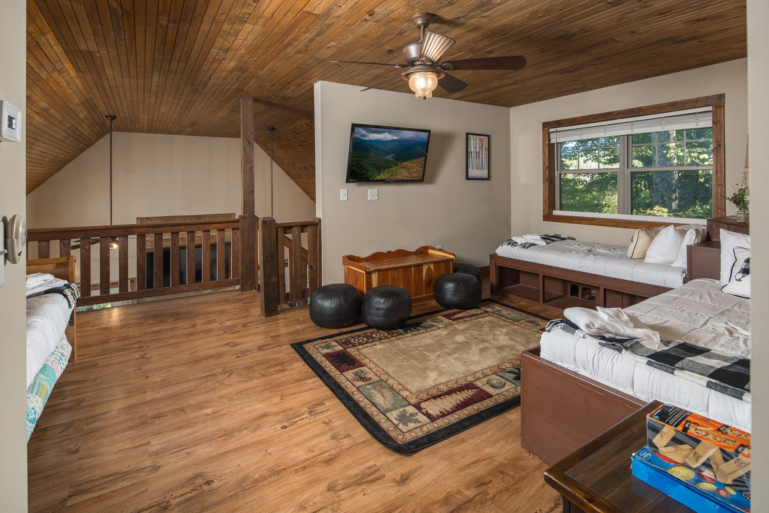 A loft bedroom with a sloped ceiling, wooden flooring, a bed, a desk, and a window with a view of trees.