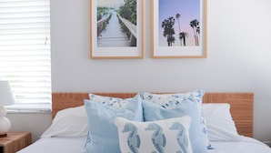 Una cama bien tendida con cabecera de madera, dos cuadros enmarcados en la pared y una mesita de noche con una lámpara.