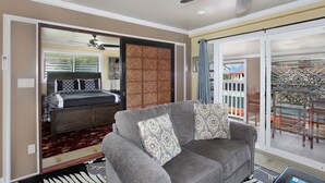 Ein Hotelzimmer mit einem Bett, einem grauen Sofa mit gemusterten Kissen, einem großen Fenster mit Blick auf einen Balkon und einem Deckenventilator.