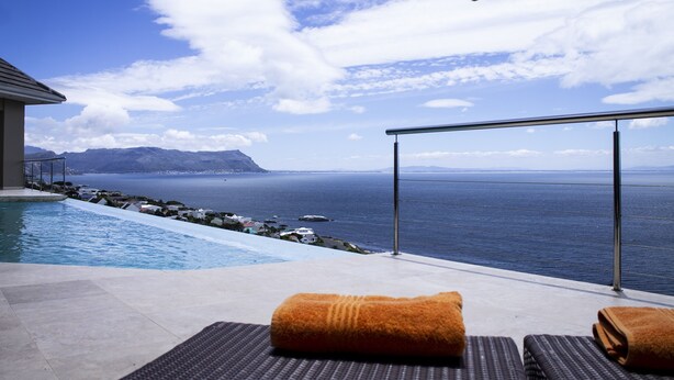 A poolside area with a view of the sea and mountains, two lounge chairs with towels, and a metal railing.